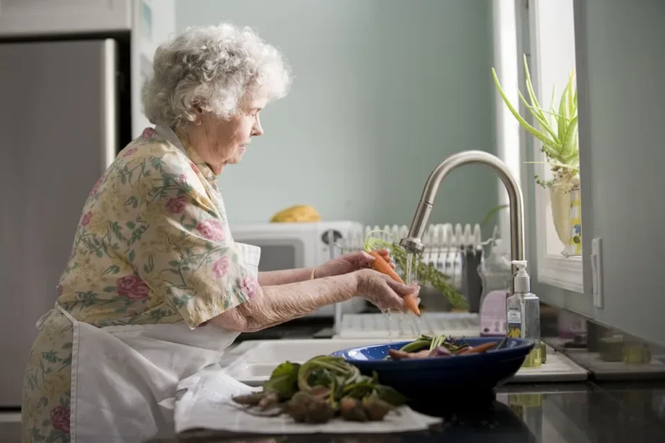 A senior woman in a floral dress washes vegetables at her kitchen sink